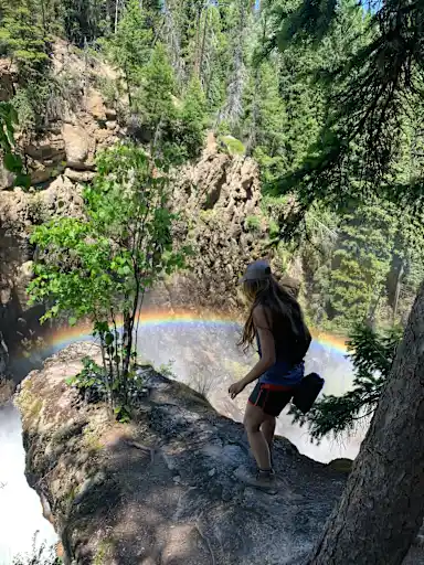 A young woman stands on a rock outcropping above a waterfall. There is a rainbow below her.